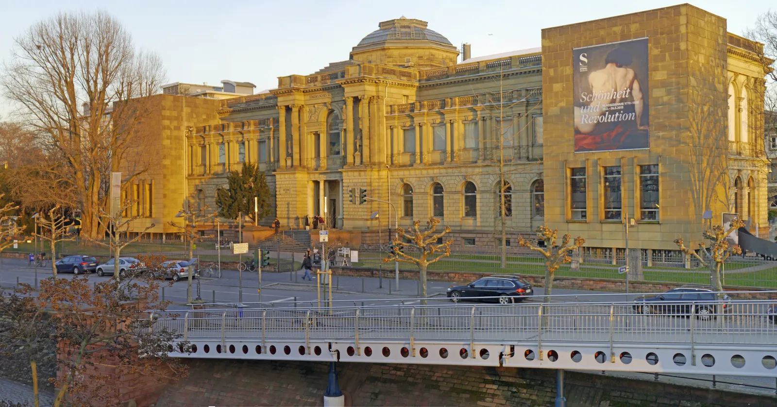 The Städel Museum's ornate sandstone facade and central dome lit gold at sunset in Frankfurt, with a pedestrian bridge crossing the Main in the foreground