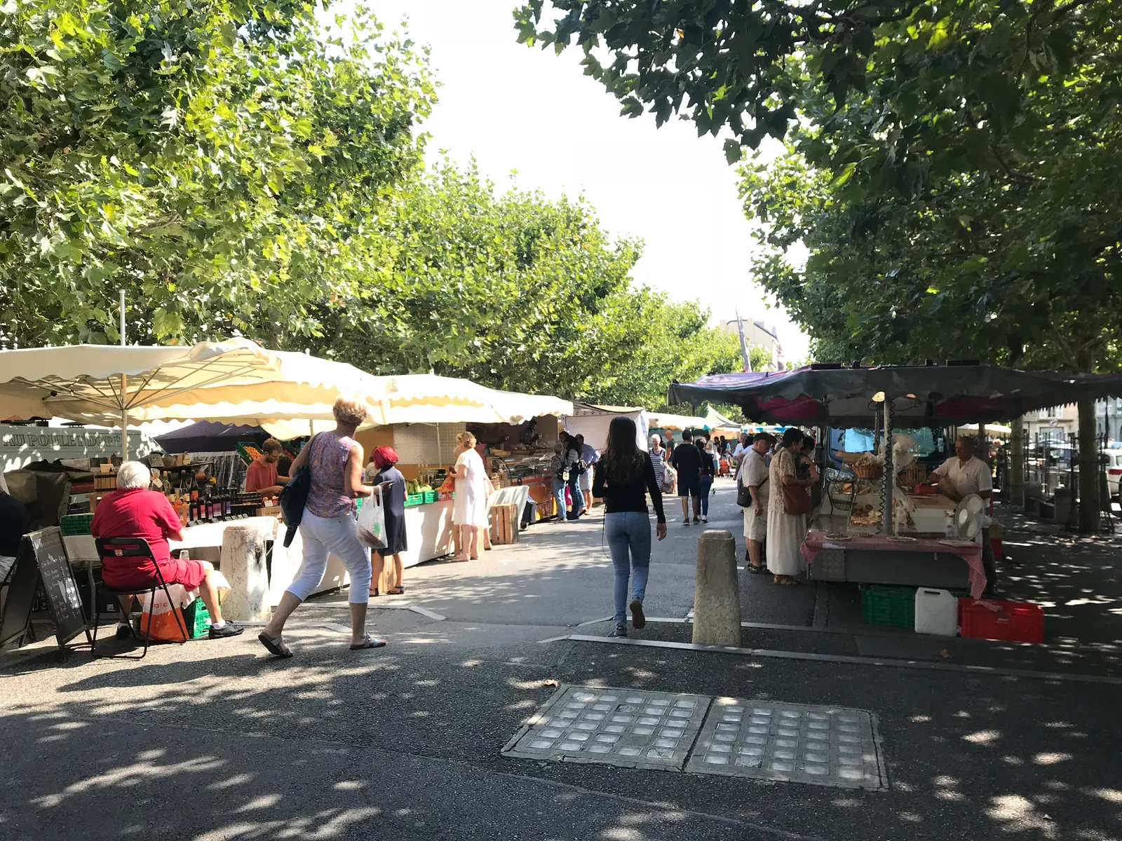 Market stalls under white awnings shaded by plane trees on Place du Marché in Carouge, shoppers moving between produce tables