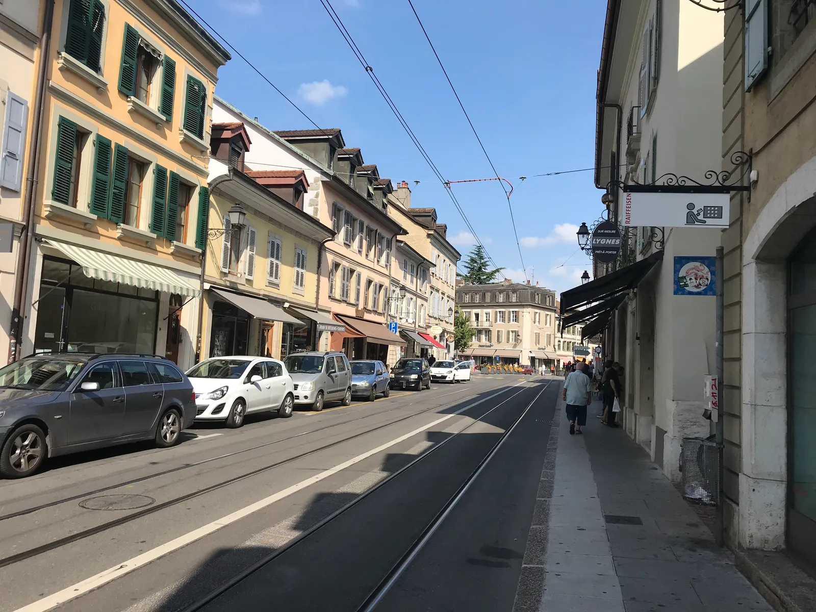 A straight Carouge street lined with two-storey shuttered houses and ground-floor shopfronts, tram rails running down the middle of the roadway