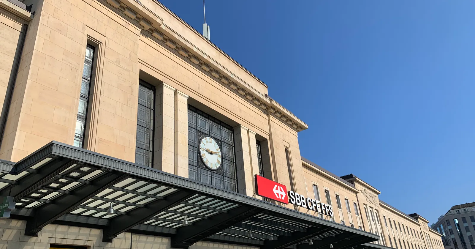 The stone frontage of Genève-Cornavin station under a blue sky, its clock set into the upper facade above a glass canopy and the railway operator's sign