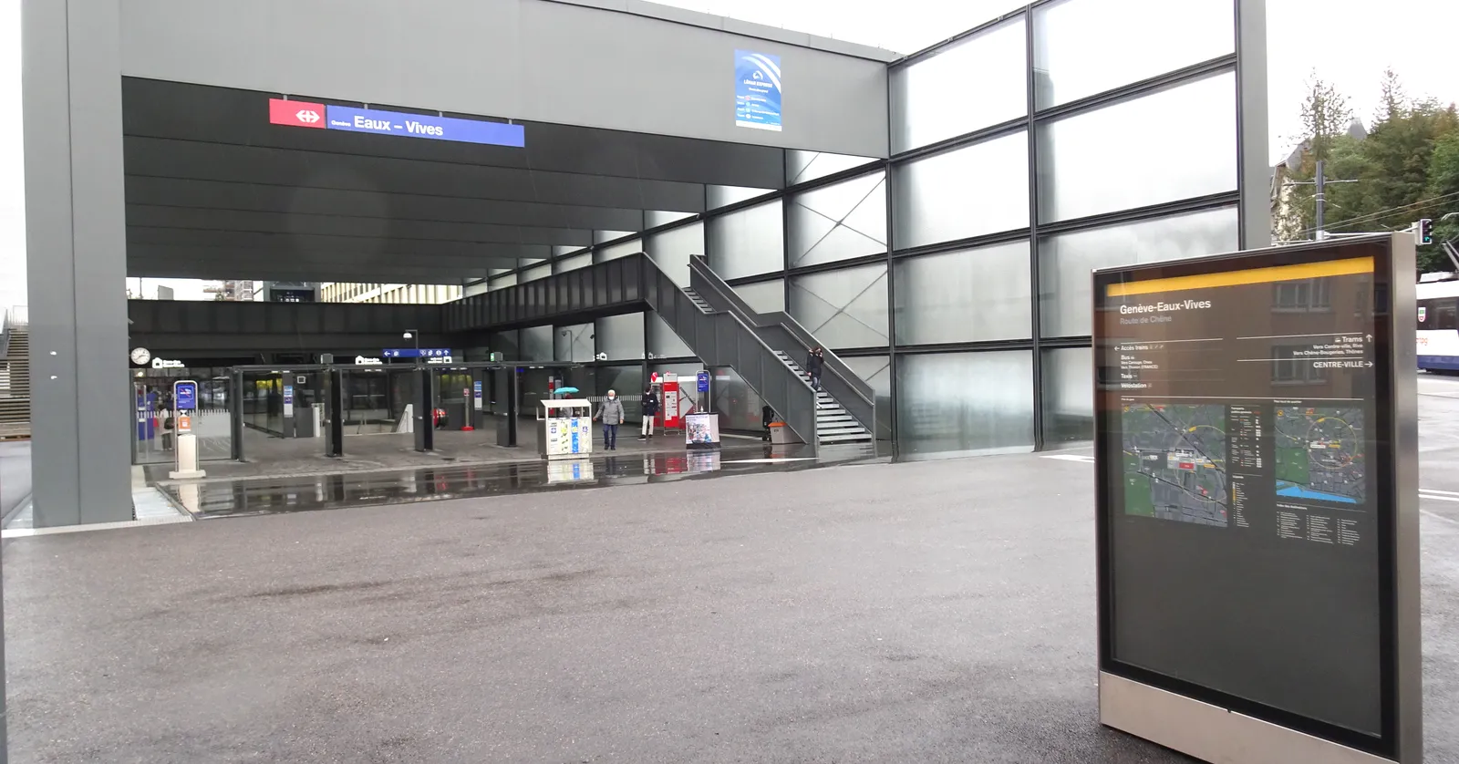 The street-level entrance hall of Genève-Eaux-Vives station, a glass-walled concourse with ticket barriers and an escalator running down to the platforms below