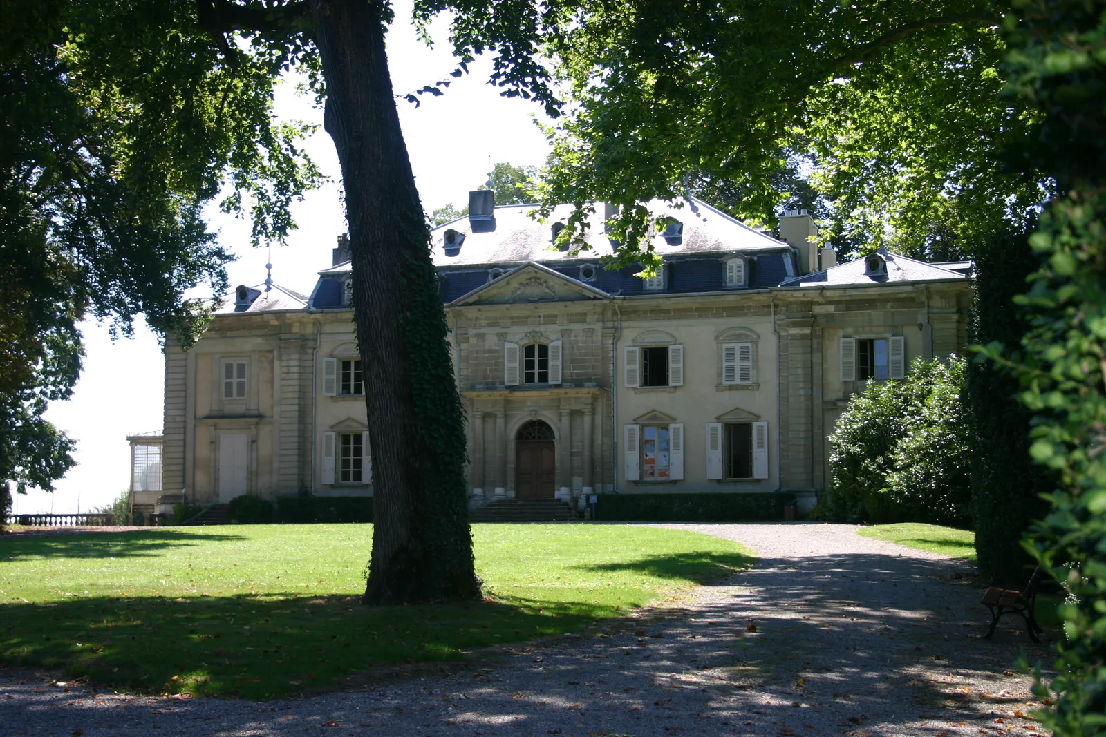 The pale classical front of Voltaire's château at Ferney seen across its lawn and gravel drive, with mature trees framing the approach