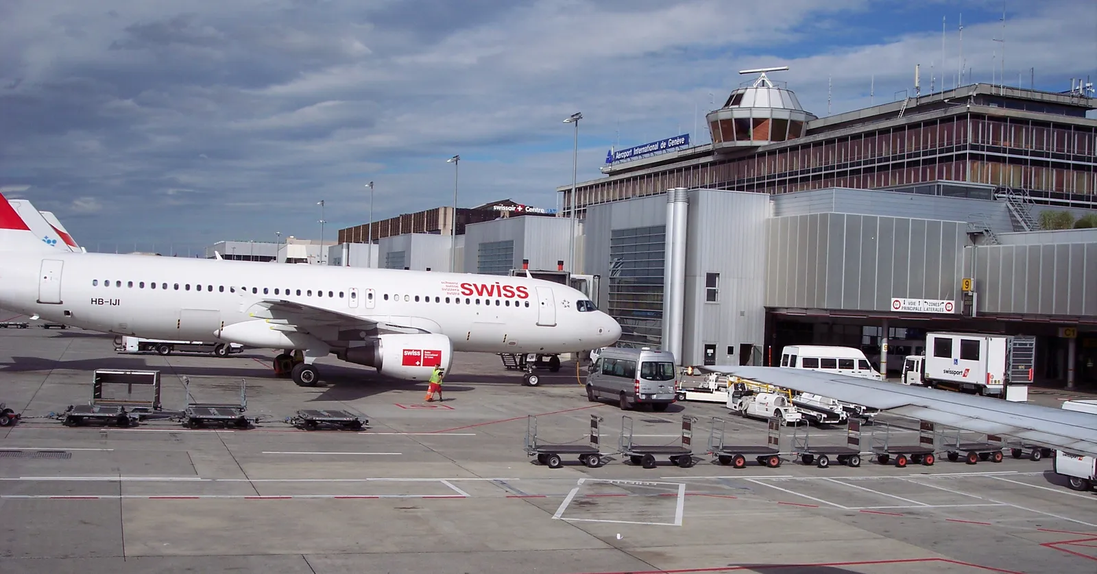 An aircraft parked at a jet bridge on the apron at Geneva Airport, the terminal building and control tower behind it under a partly clouded sky