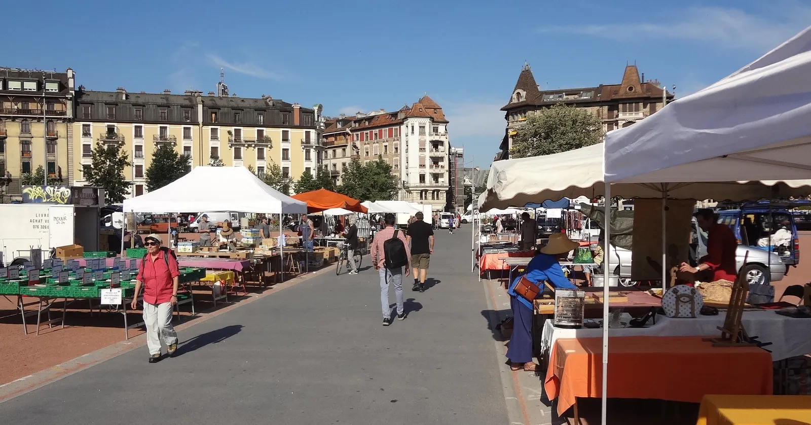Flea market stalls under white and orange awnings spread across the open Plaine de Plainpalais in Geneva, browsers walking between them with apartment blocks behind