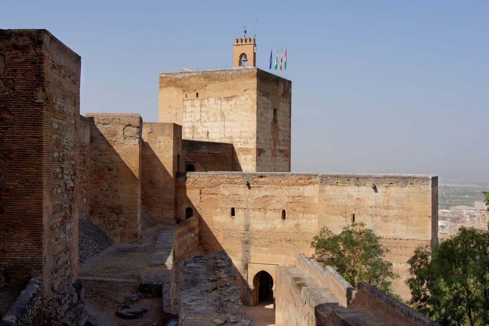 The Alcazaba fortress at the western tip of the Alhambra, sandstone ramparts and a square watchtower flying flags, with Granada's rooftops visible in the haze beyond