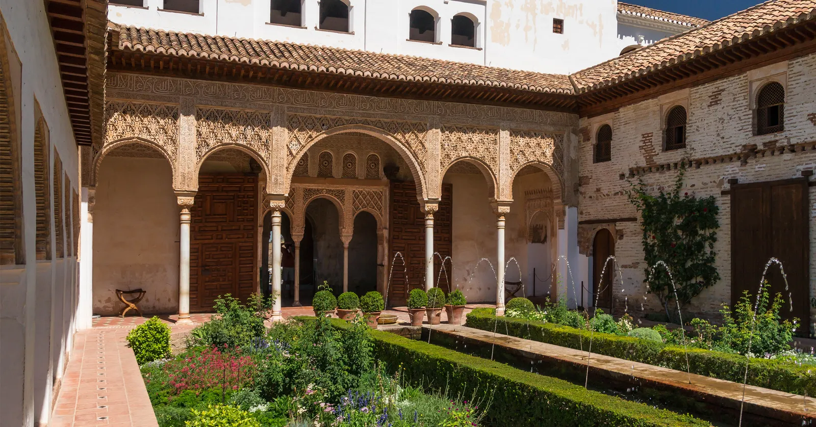 The Patio de la Acequia in the Generalife, Granada, a long garden courtyard with a water channel between flowerbeds and arching fountain jets, an arcaded pavilion with carved plasterwork at the far end