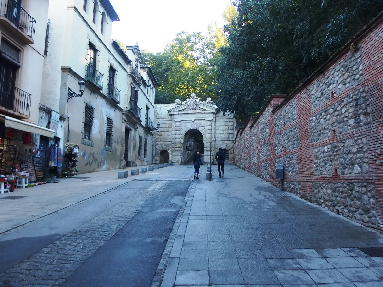 The Puerta de las Granadas at the top of Cuesta de Gomérez in Granada, a Renaissance stone gateway leading into trees, with a brick-and-rubble wall along one side of the sloping street