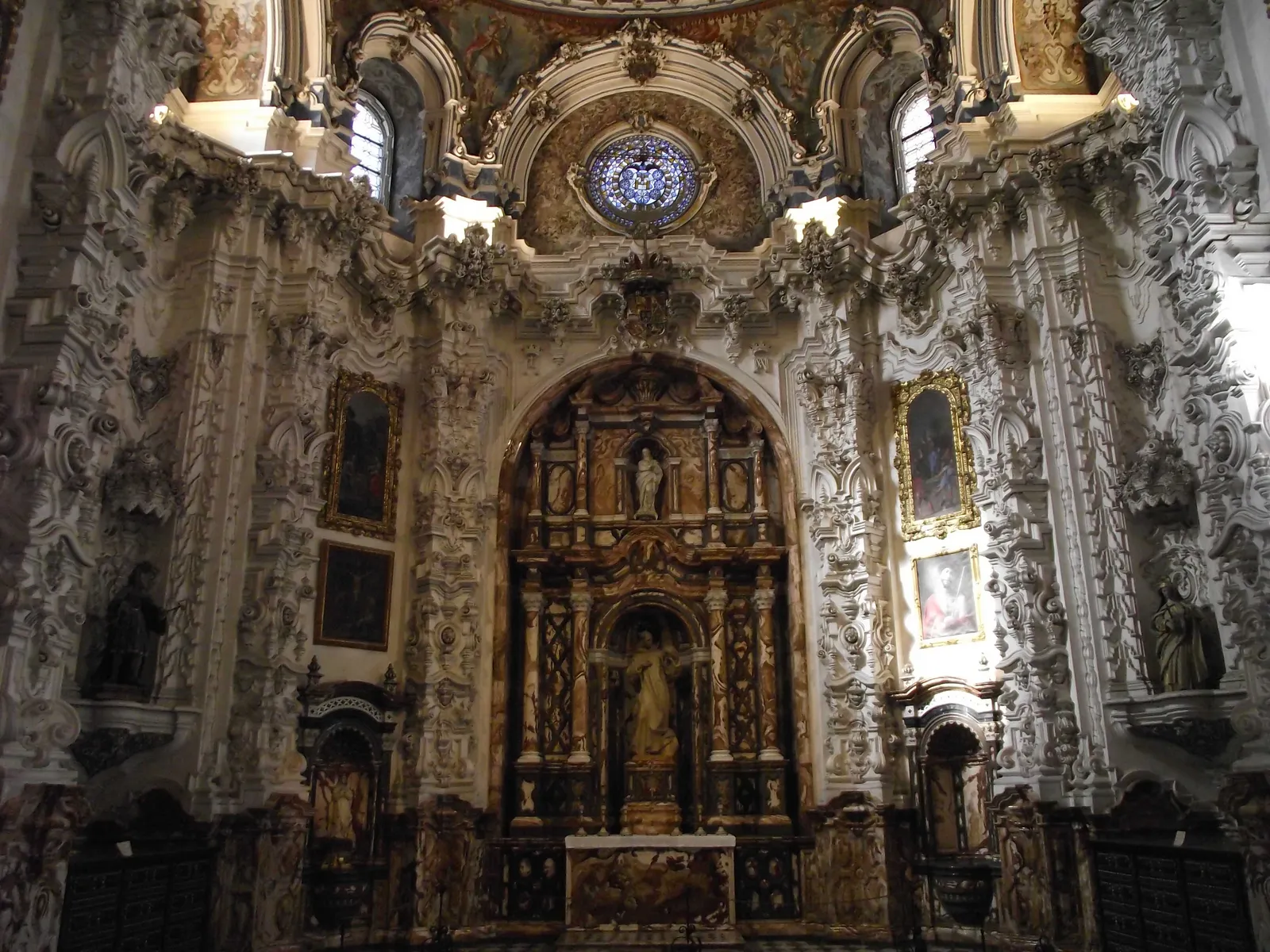 The sacristy of the Cartuja monastery in Granada, white stucco carved into dense Baroque mouldings around a gilded altarpiece, dark painted panels on the walls