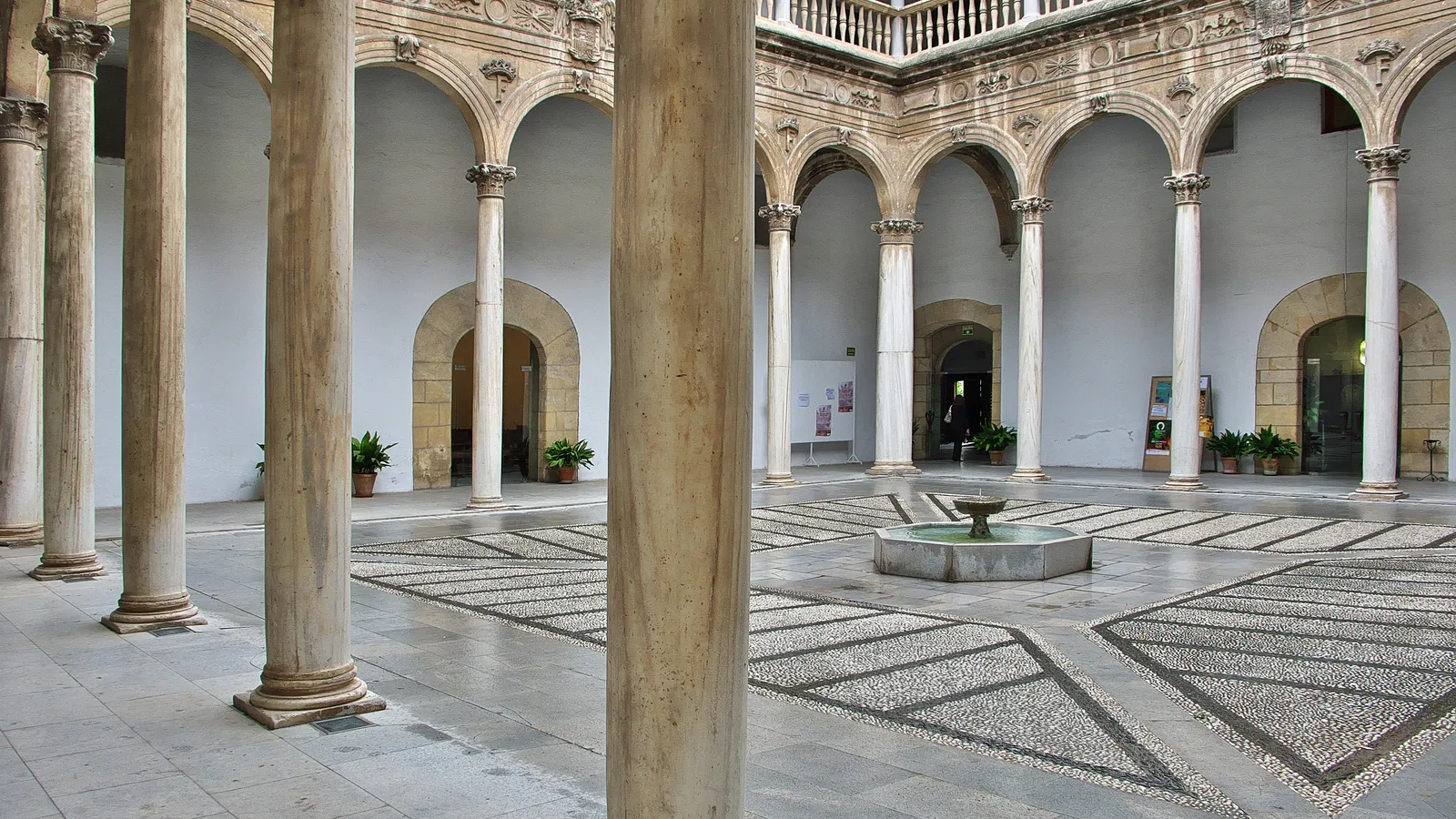 The Patio de los Mármoles inside Granada's Hospital Real, a two-storey Renaissance arcade of stone columns around a pebble-mosaic courtyard with a low fountain in the middle