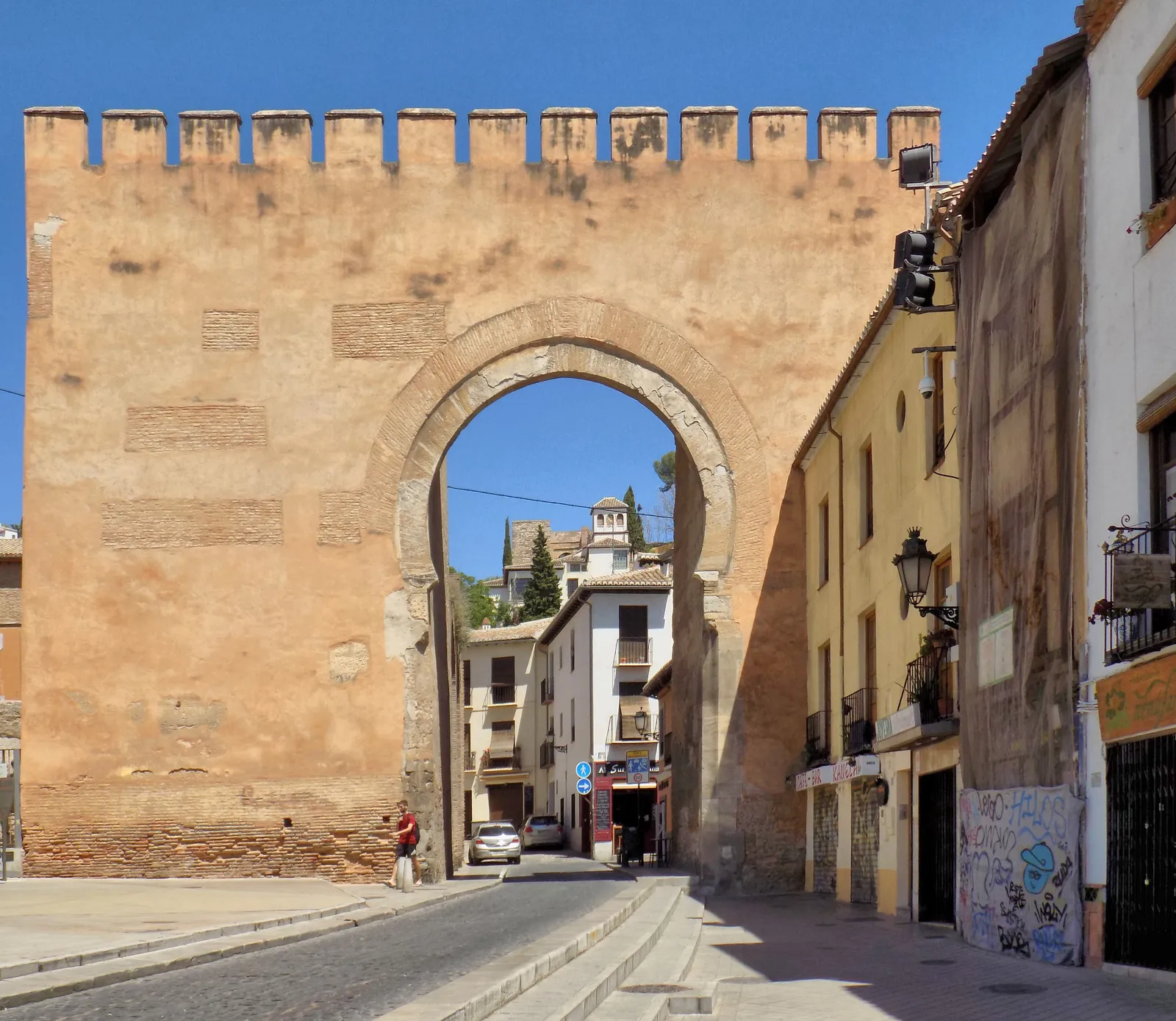 The Puerta de Elvira in Granada, a tall crenellated brick-and-rubble gate with a horseshoe arch, a narrow street and houses of the Albaicín visible through it