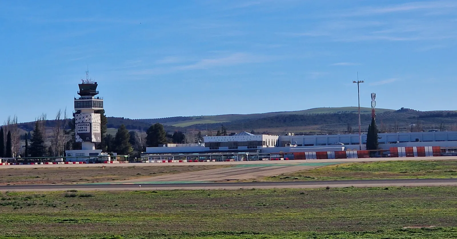 Federico García Lorca Granada-Jaén Airport seen across open fields, the control tower and the low terminal buildings under a blue sky with hills behind