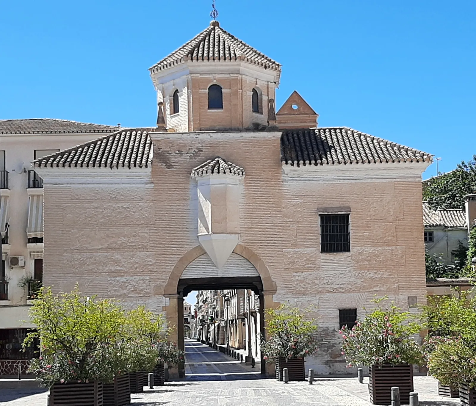 The Puerta de Granada in Santa Fe, a pale gatehouse with a tiled roof and a small domed lantern on top, its arch opening onto a straight pedestrian street