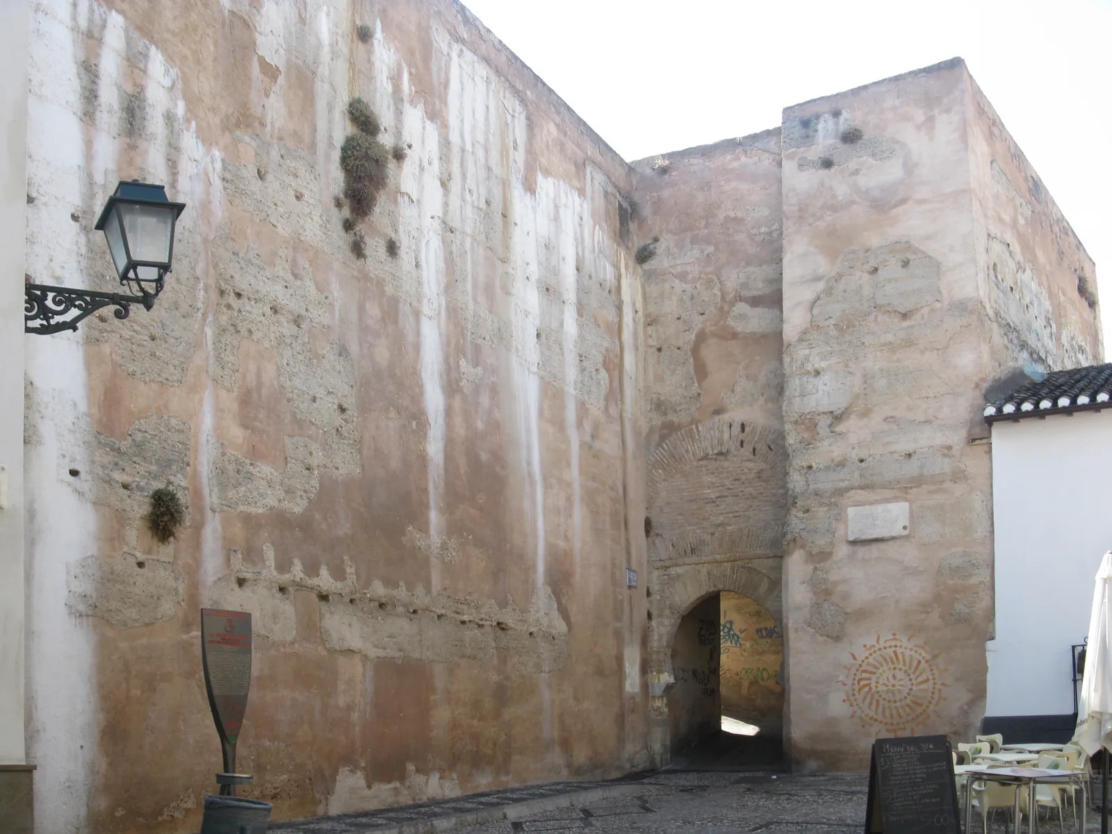 The Arco de las Pesas in Granada's upper Albaicín, a weathered stone gate tower in the old city wall with a low arched passage through it and café tables beside it