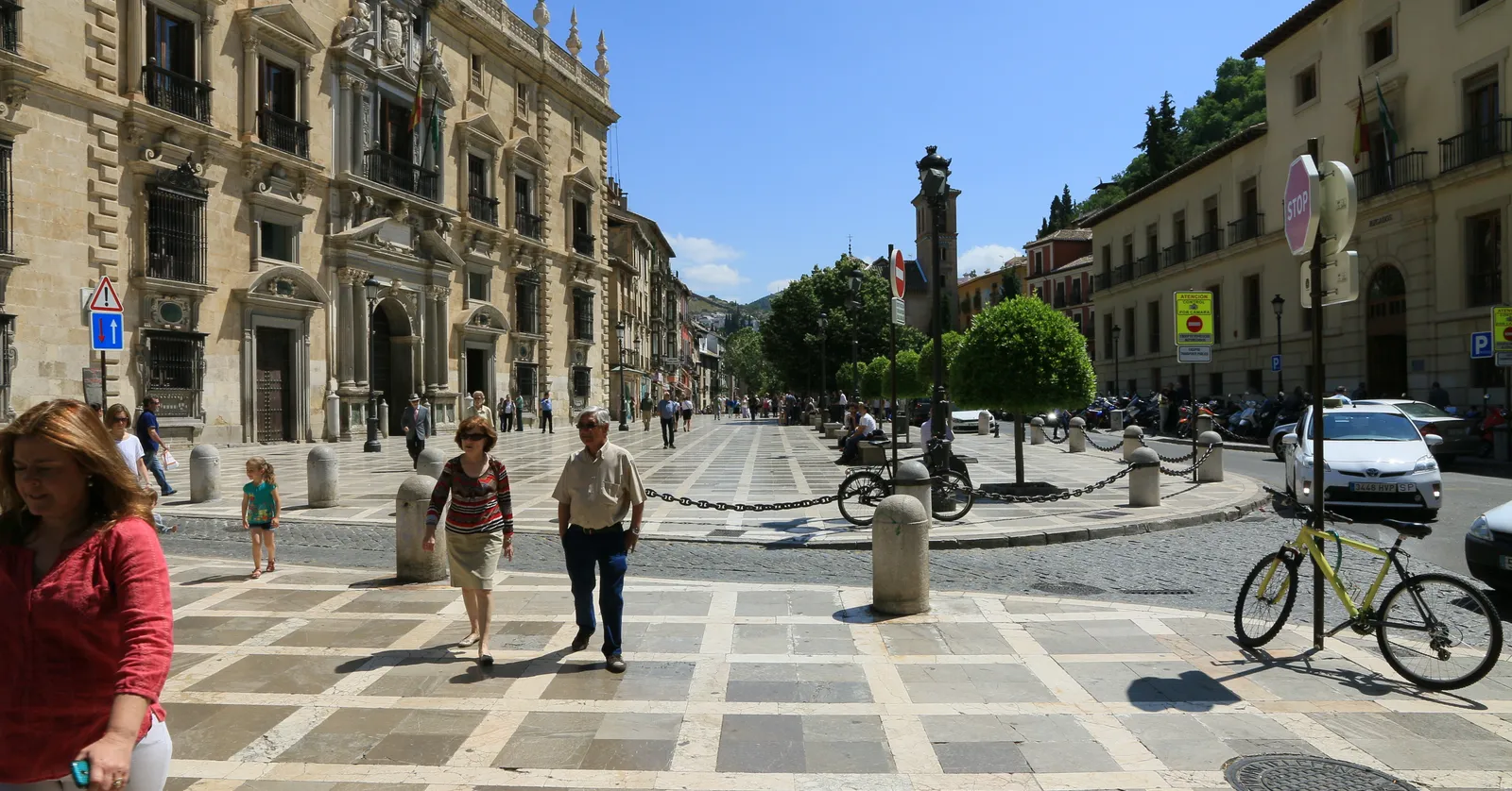 Plaza Nueva in Granada on a sunny day, the carved stone front of the Real Chancillería on the left, a paved square with bollards and cyclists, and the wooded Alhambra hill rising behind the far end