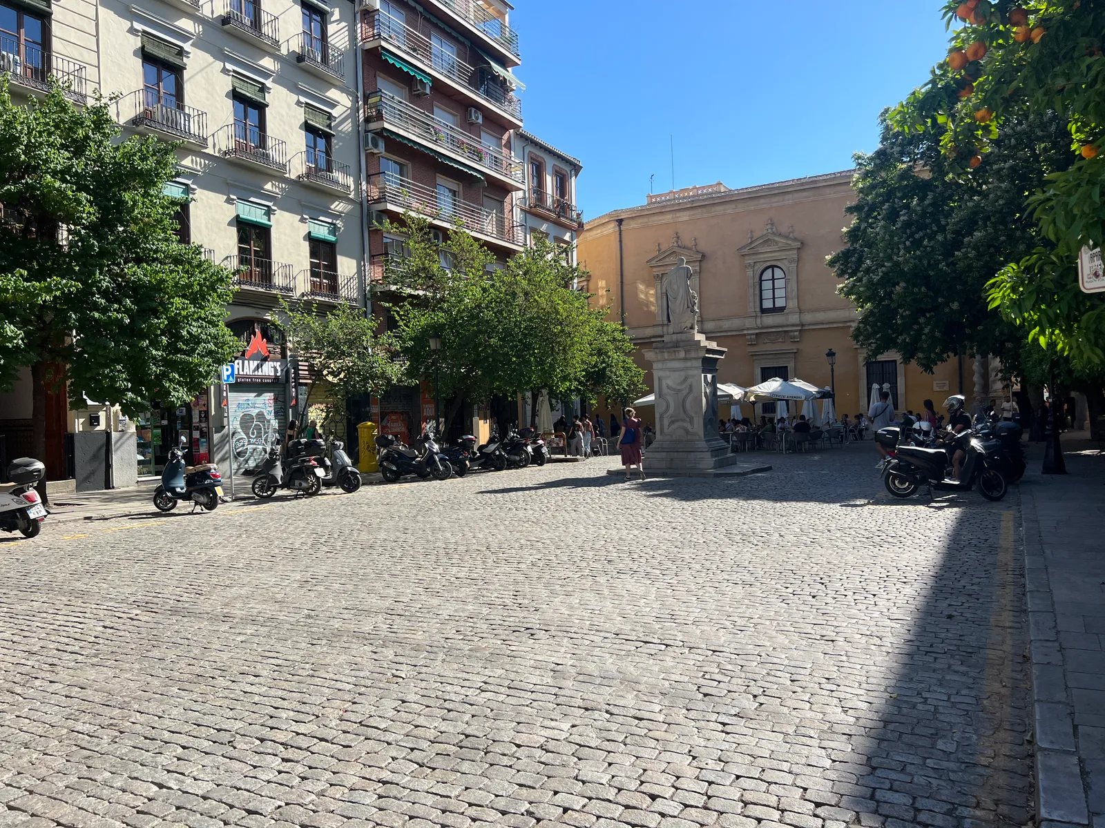 Plaza de la Universidad in Granada, a cobbled square lined with orange trees and parked scooters, a monument on a tall pedestal in the middle and an ochre Baroque facade behind