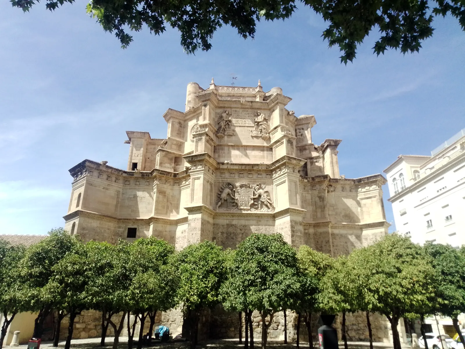 The stone east end of the Monastery of San Jerónimo church in Granada, carved coats of arms high on its walls, seen over a row of orange trees