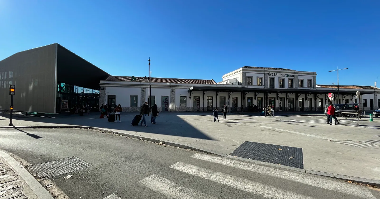 The forecourt of Granada railway station on a sunny day, the low cream-coloured 19th-century station building with a modern dark extension at one end and travellers wheeling suitcases across the square