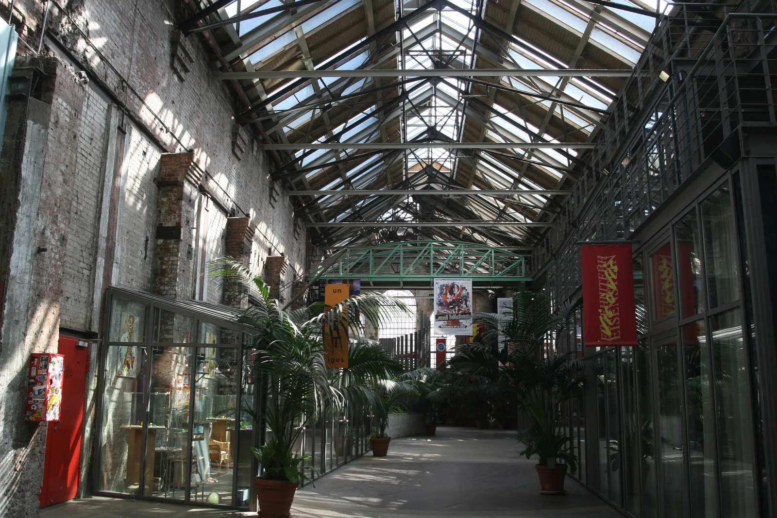 The glass-roofed interior arcade of the Zeisehallen in Hamburg-Ottensen, a former factory hall with exposed brick walls, potted palms and cinema posters along one side