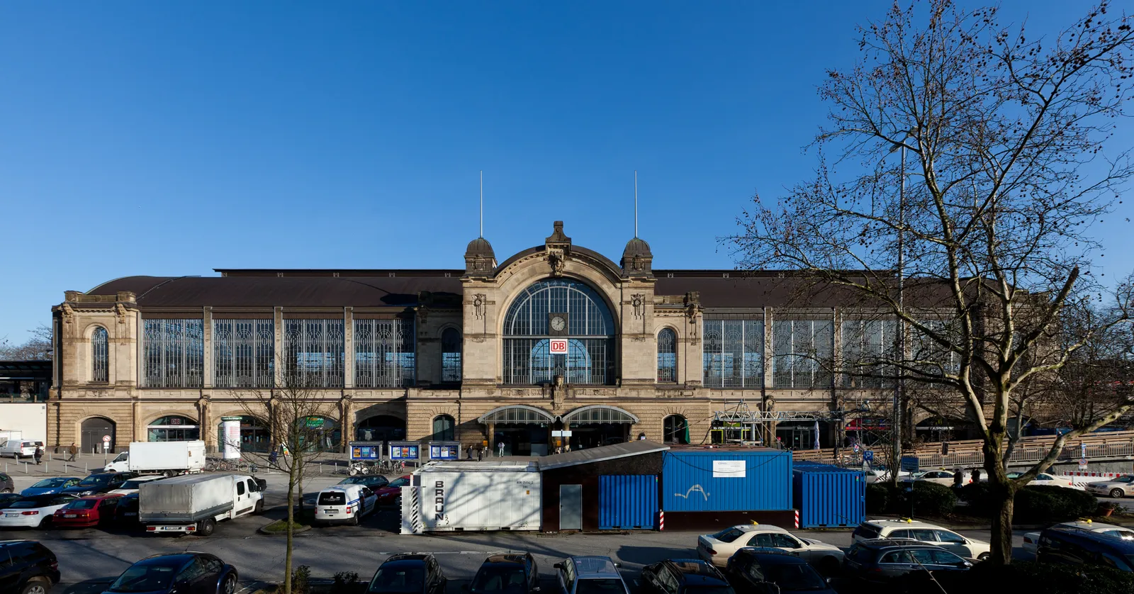 The long sandstone front of Hamburg Dammtor station under a blue sky, with its arched central gable and rows of tall glazed windows lighting the train hall behind