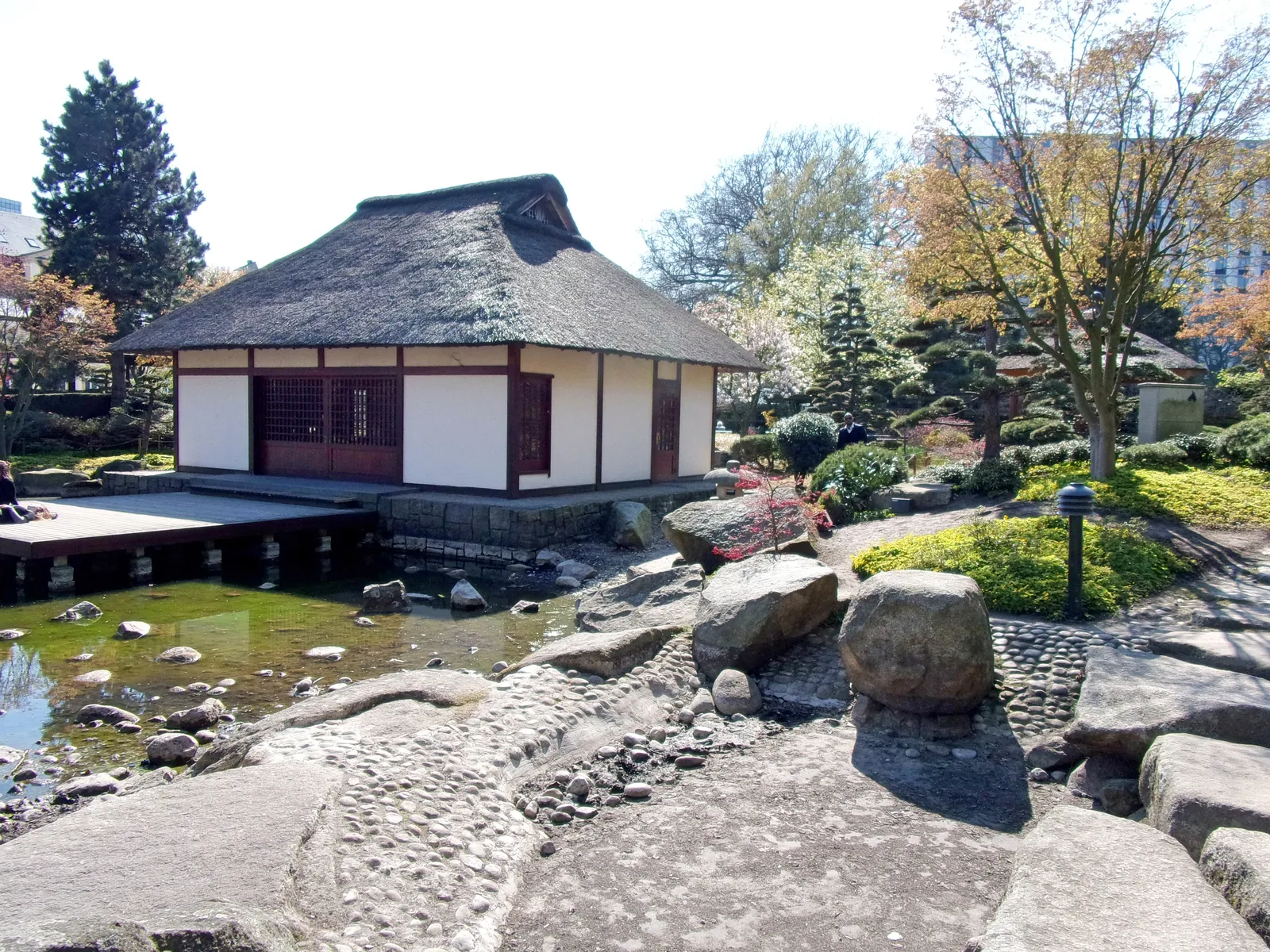 A thatched wooden tea house at the edge of a pond in the Japanese garden at Planten un Blomen in Hamburg, with clipped shrubs and stepping stones in the foreground