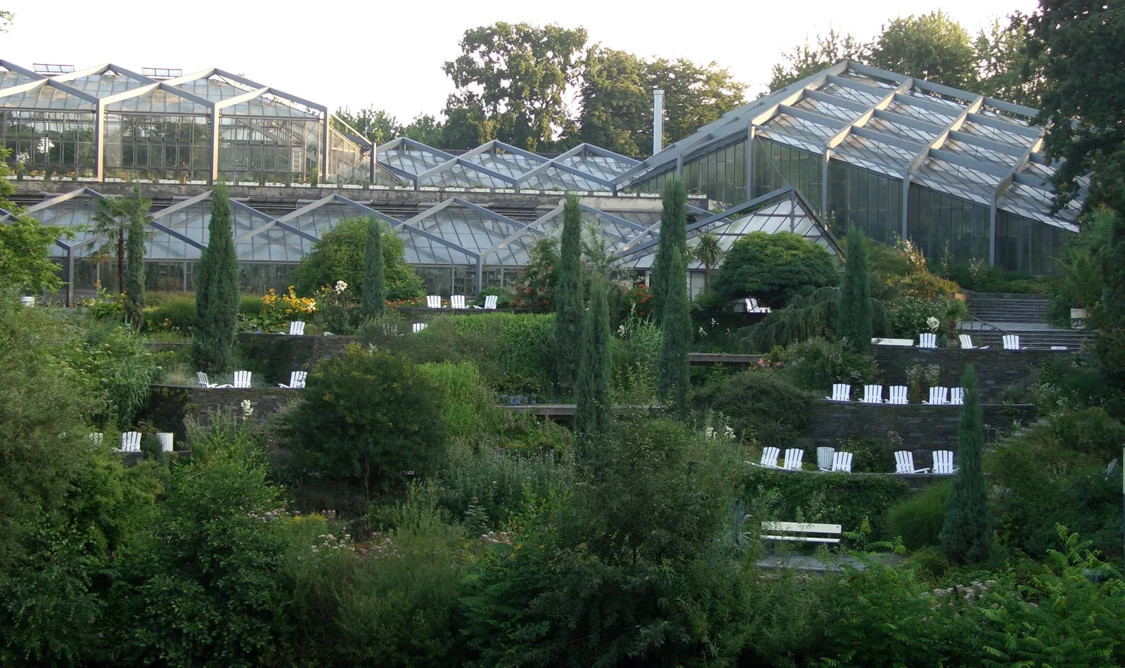 The long ridged glasshouses of the tropical greenhouses at Planten un Blomen in Hamburg, seen across terraced garden beds and white deckchairs on the slope below