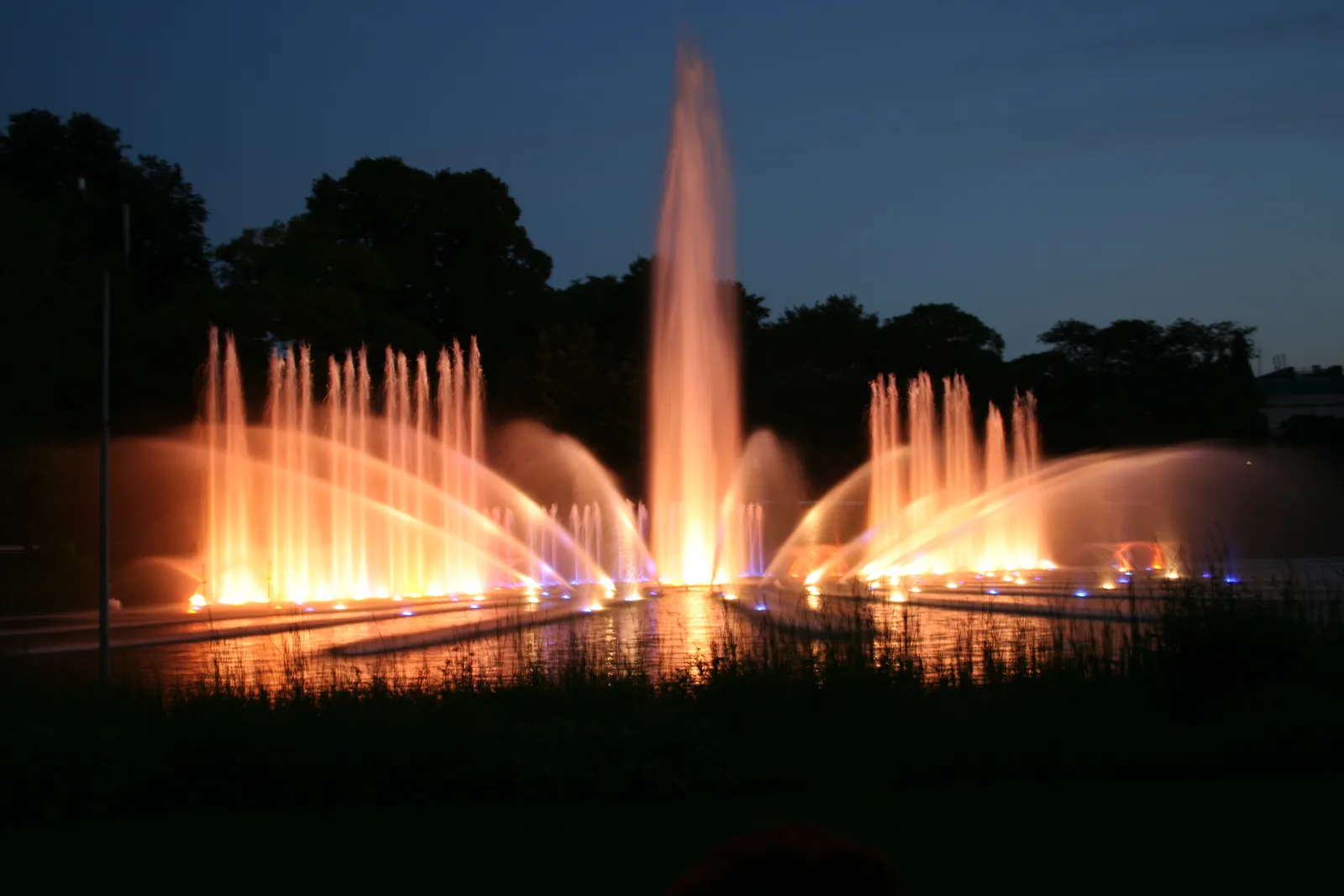 Jets of water lit orange and blue rising from the lake at Planten un Blomen in Hamburg after dark, reflected in the still water below
