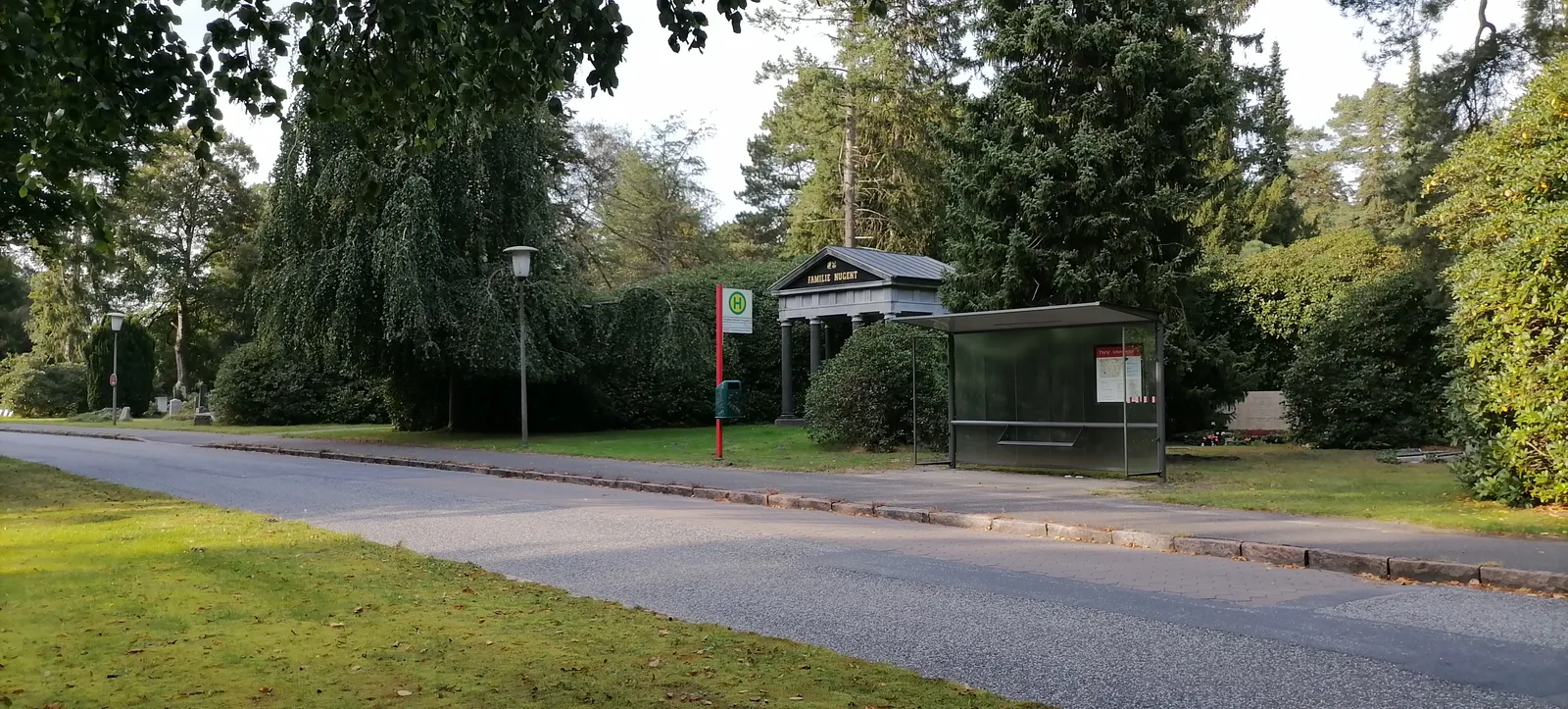 A glass bus shelter and timetable post beside a paved road running between trees inside Ohlsdorf Cemetery in Hamburg