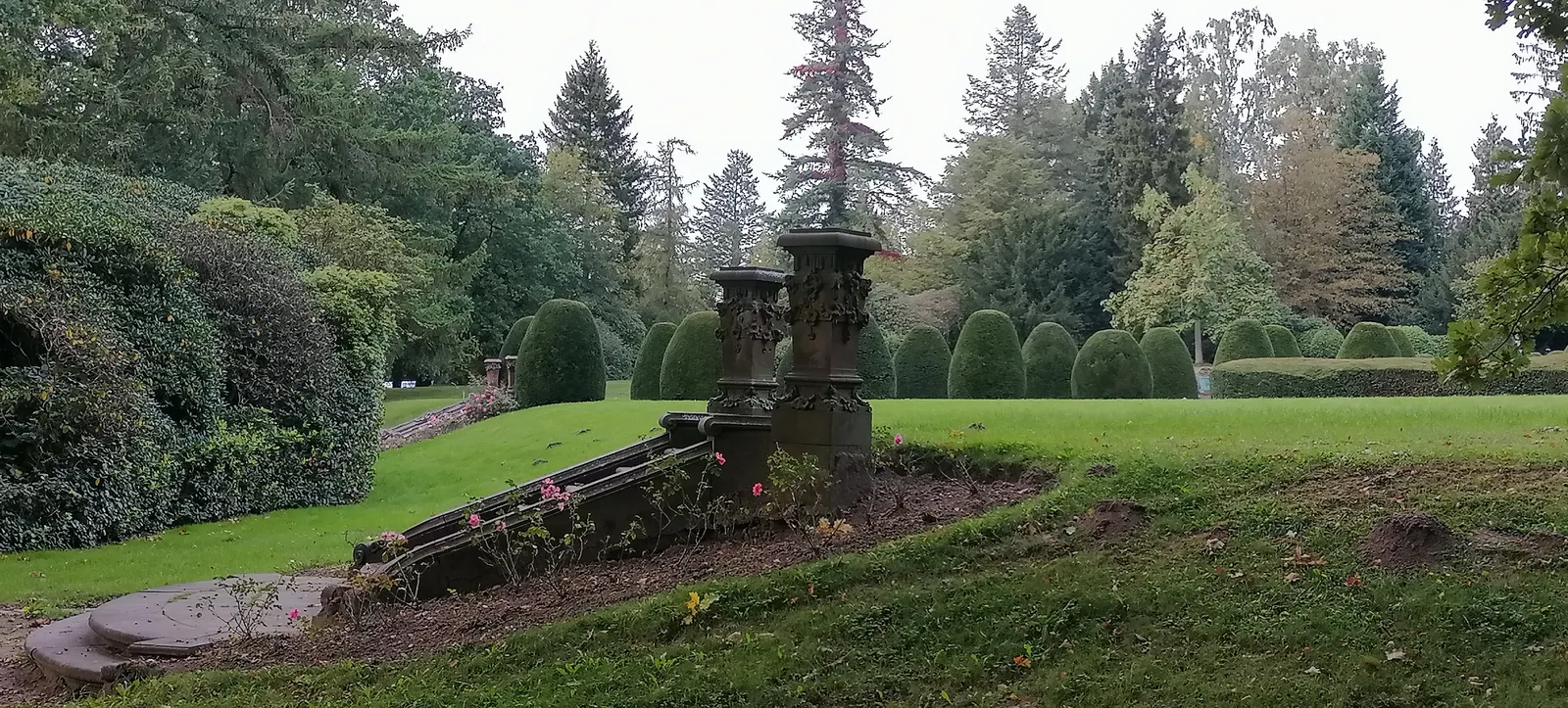 A wide lawn at Ohlsdorf Cemetery in Hamburg with clipped hedges, mature conifers and a stone urn on a plinth in the foreground