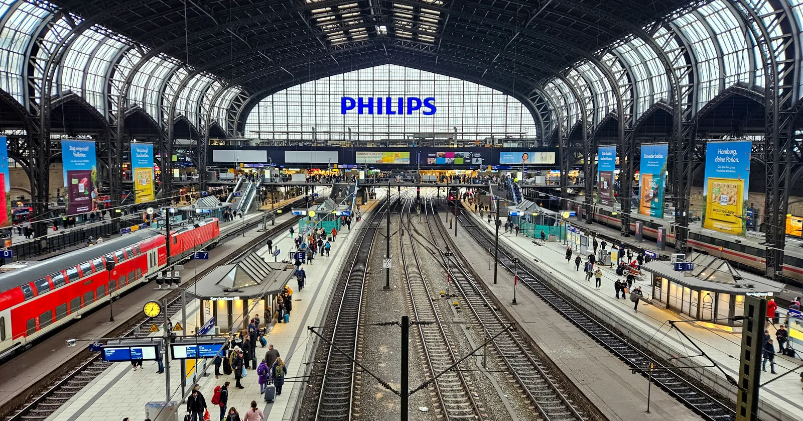 The arched iron-and-glass roof of Hamburg Hauptbahnhof seen from the overhead bridge, with red regional trains at the platforms below and travellers crossing the concourse
