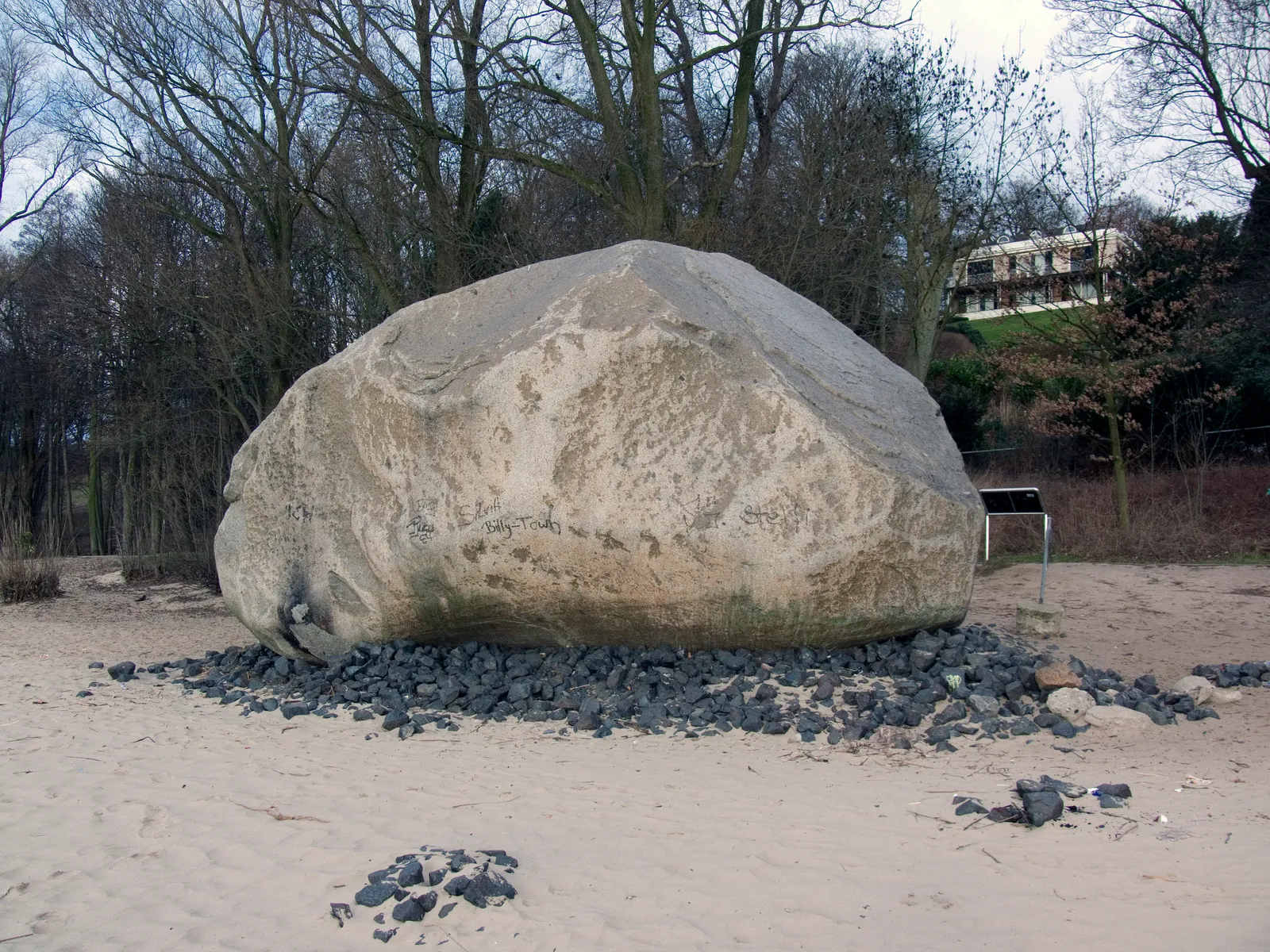 A very large pale granite boulder sitting on the sand of the Elbe beach at Övelgönne in Hamburg, ringed by dark stones with bare trees behind
