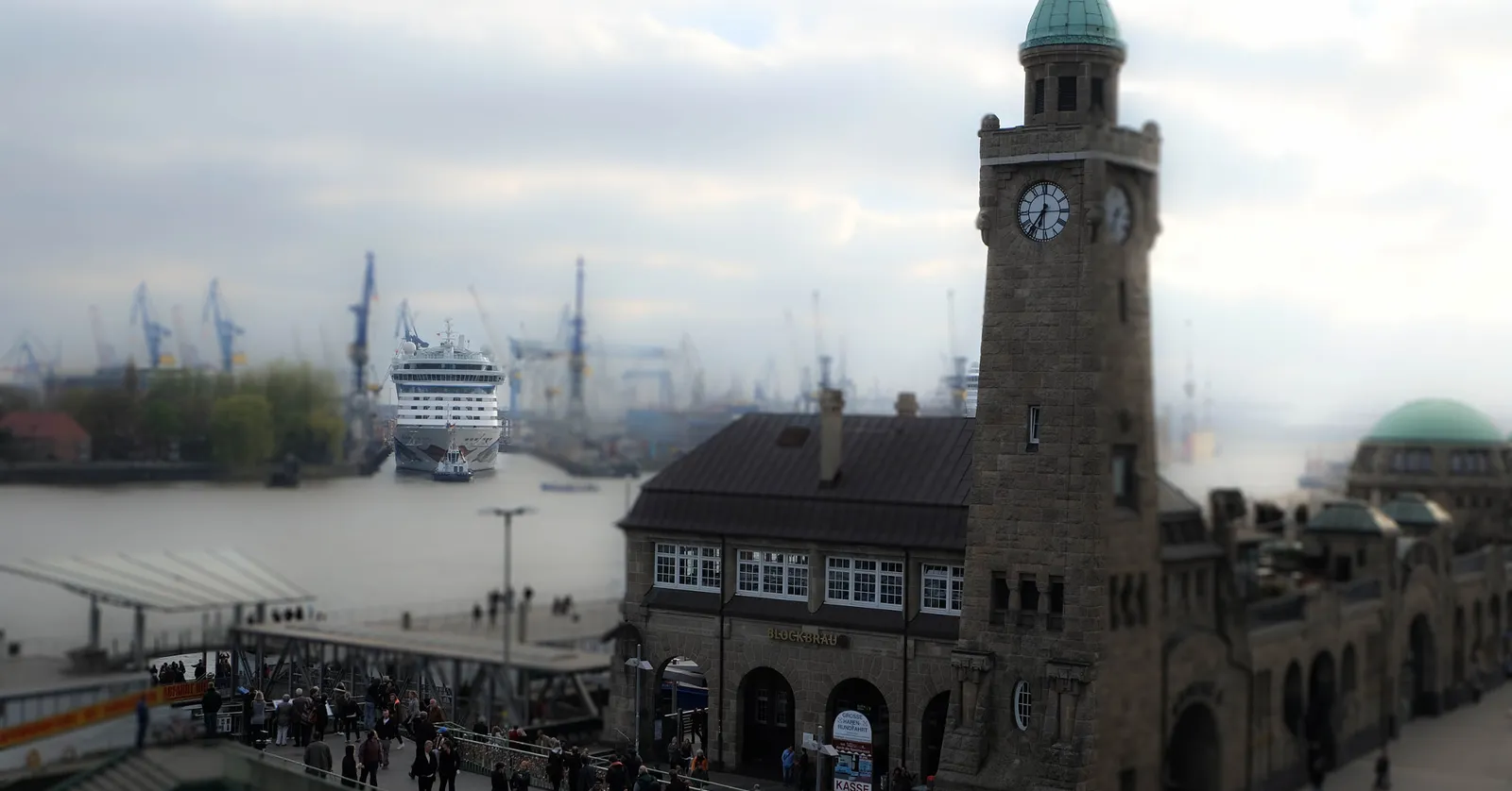 The stone clock and tide-gauge tower of Hamburg's Landungsbrücken above the pier building, with a cruise ship and harbour cranes across the water behind it