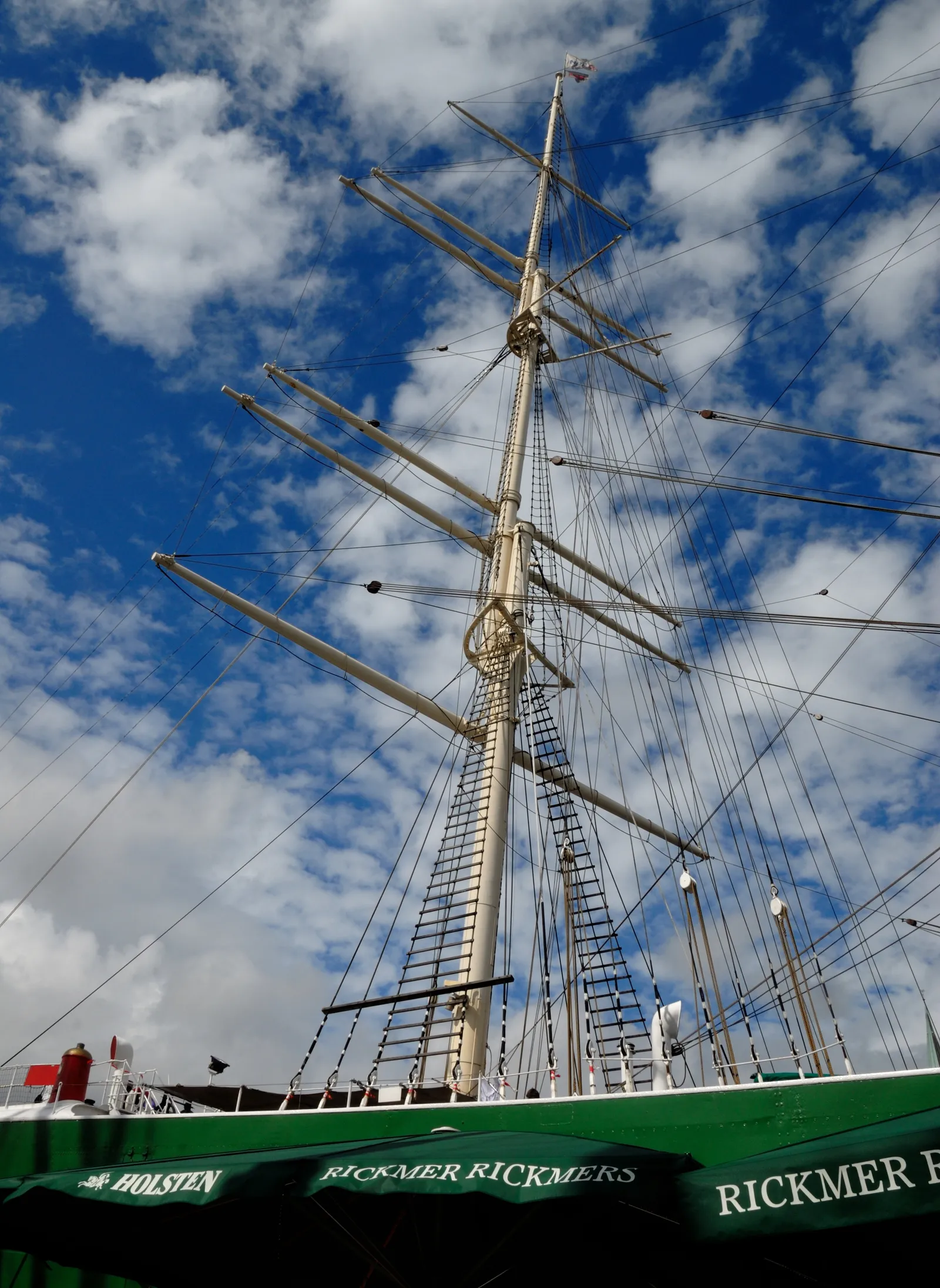 The masts and rigging of the three-masted barque Rickmer Rickmers against a cloudy sky, with the ship's green hull and name lettering along the bottom of the frame