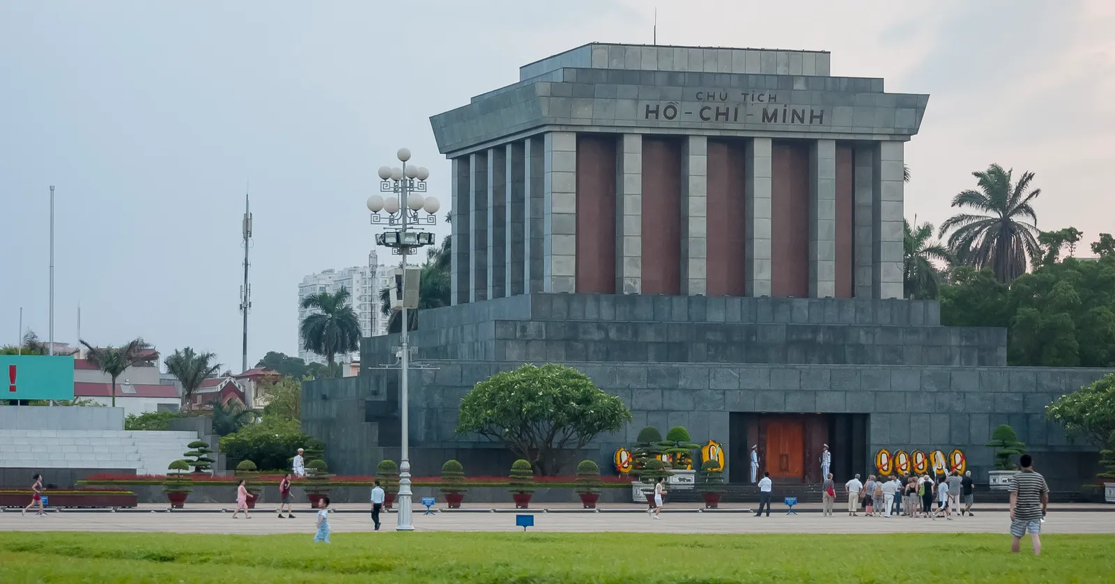 The grey granite Ho Chi Minh Mausoleum seen across the mown lawns of Ba Dinh Square in Hanoi, its name carved across the front above a colonnade, with people walking along the road in front