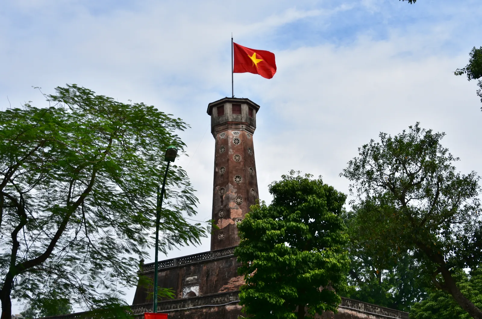 The brick Flag Tower of Hanoi rising above its walled base between trees, an octagonal column with small windows and the red Vietnamese flag flying from the observation turret on top