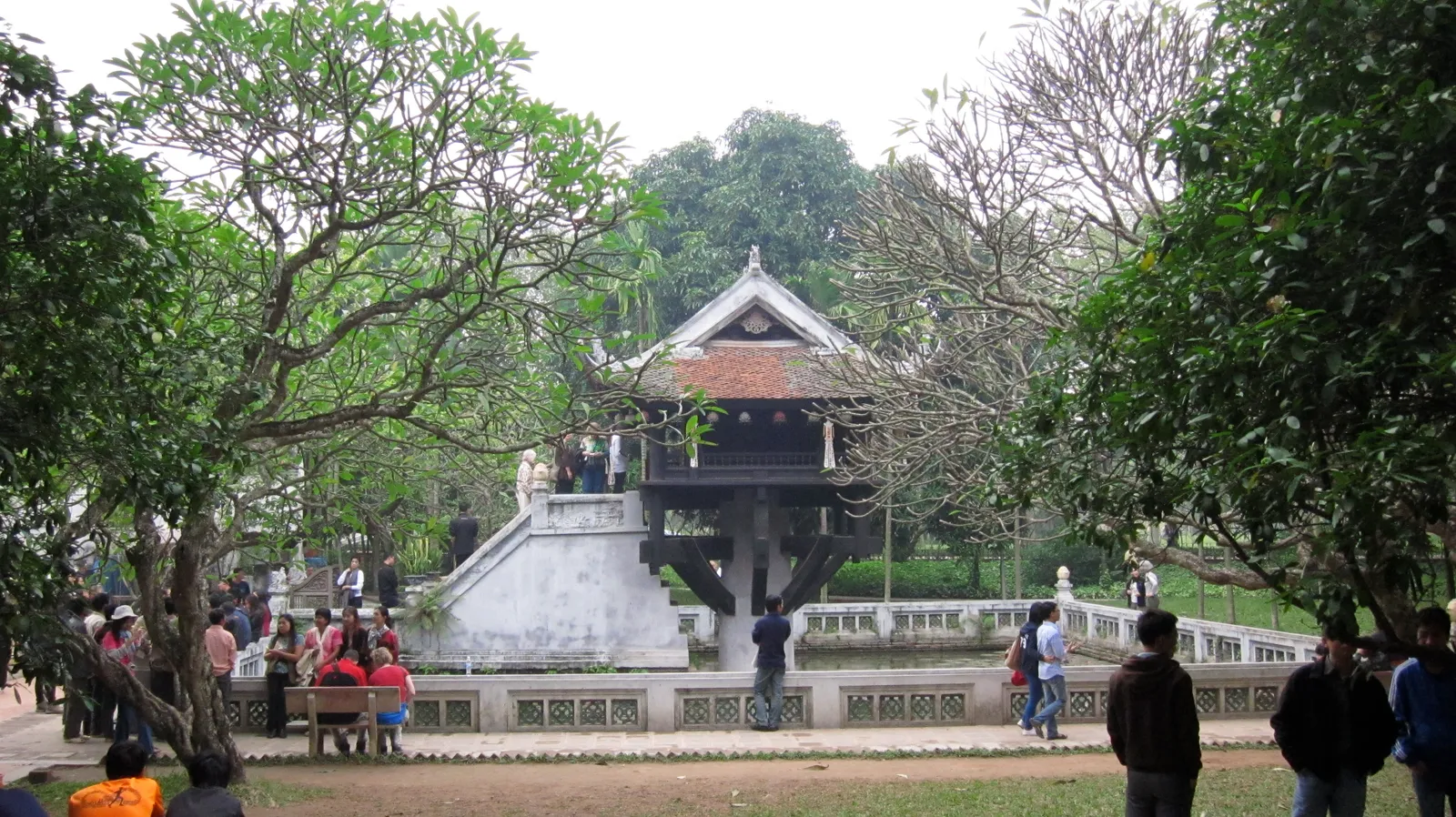 The One Pillar Pagoda in Hanoi, a small dark wooden shrine with a tiled roof raised on a single stone column above a square pond, reached by a narrow flight of steps, with visitors and frangipani trees around it