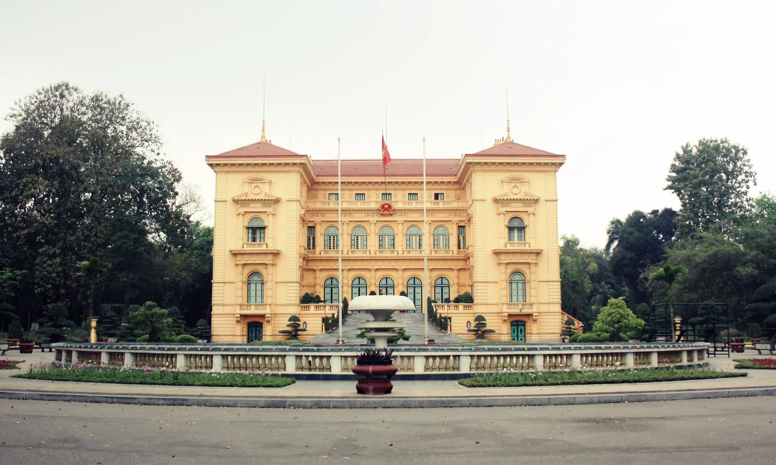 The Presidential Palace in Hanoi, a symmetrical mustard-yellow colonial mansion with a red-tiled roof, a flag over the central balcony and a fountain on the drive in front of it