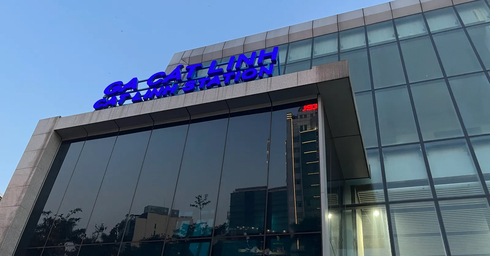 The glass-fronted entrance building of Cat Linh metro station in Hanoi at dusk, with GA CAT LINH and the English station name lit in blue letters along the roofline