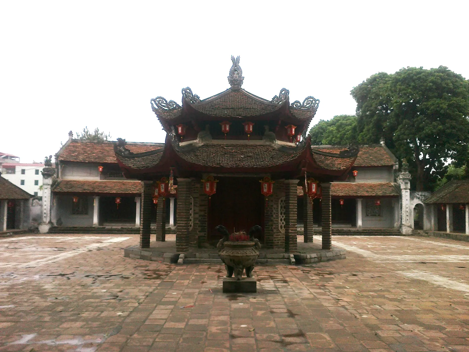 The two-tier octagonal pavilion in the courtyard of Lang Pagoda in Hanoi, dark timber and curved tiled roofs hung with red lanterns, with a stone incense burner on the brick paving in front