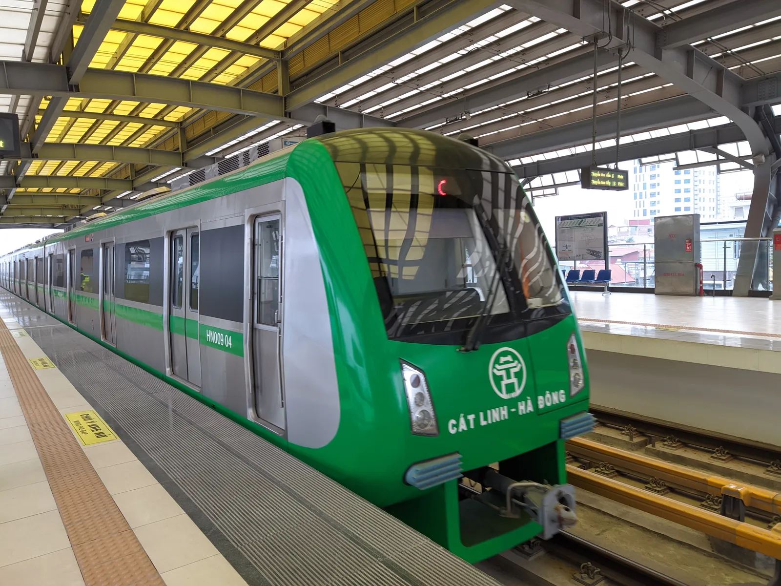 A bright green four-carriage metro train lettered Cat Linh - Ha Dong drawing into an elevated platform in Hanoi under a yellow-lit steel canopy