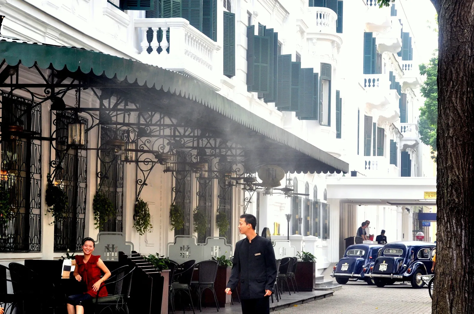 The white colonnaded frontage of the Metropole hotel in Hanoi with its green striped awning over the terrace tables, a vintage black car parked at the kerb beyond