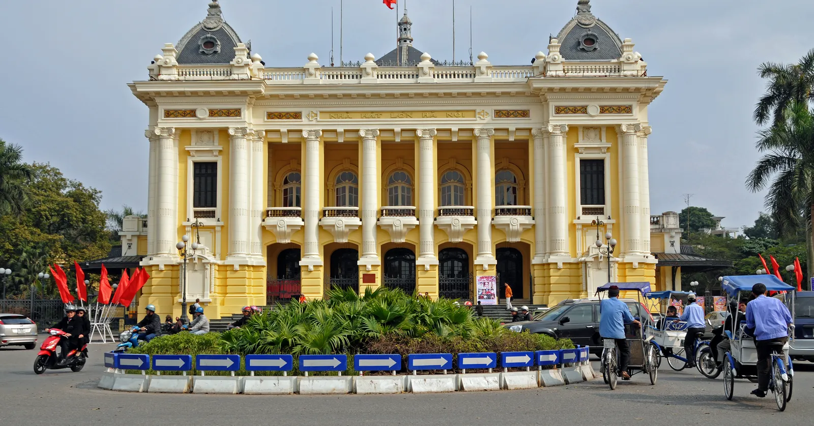 The Hanoi Opera House, a pale yellow French neoclassical building with a columned upper loggia, twin domed pavilions and a red flag over the centre, seen head-on across the roundabout in front of it
