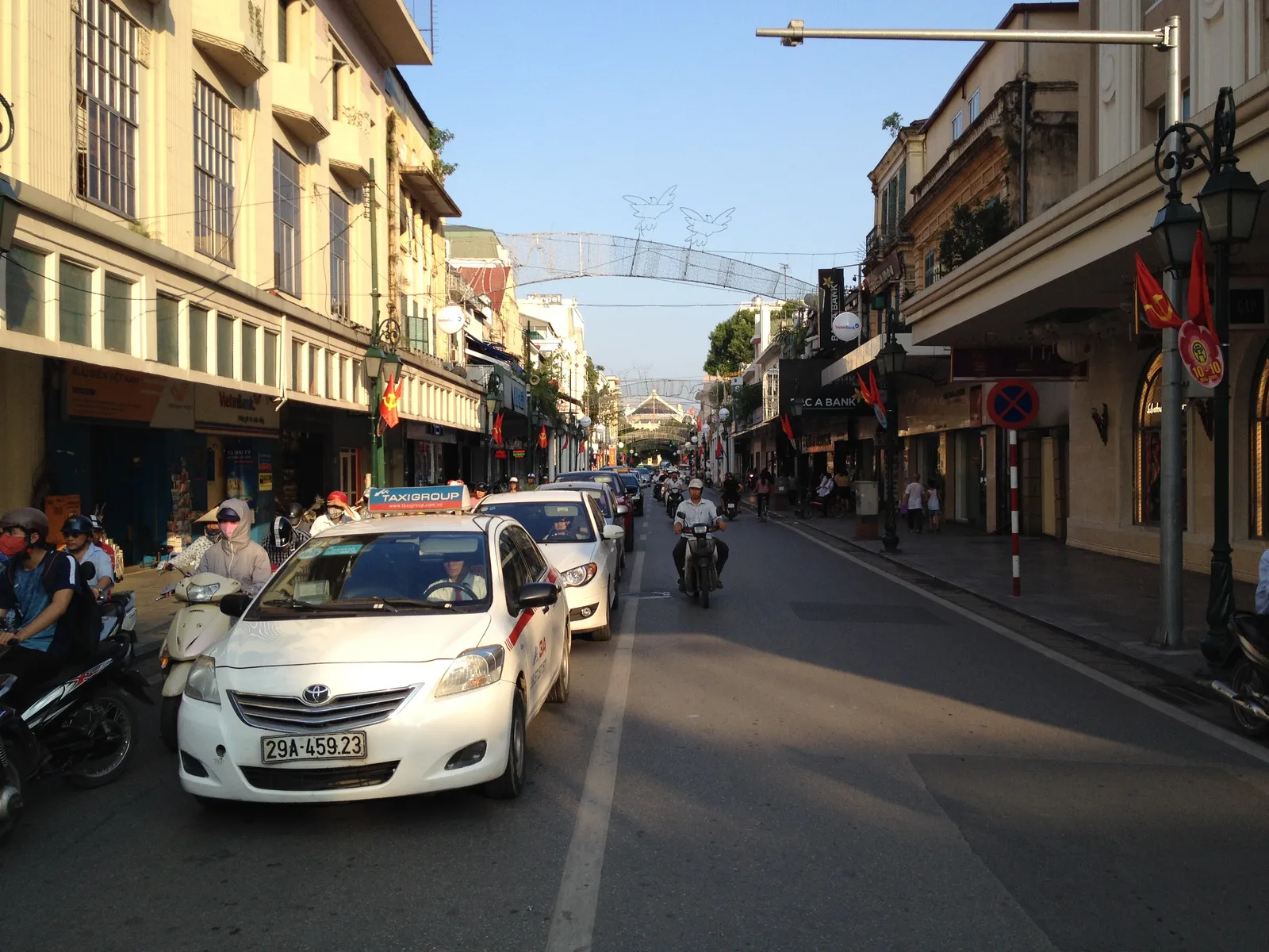 Trang Tien street in Hanoi in late afternoon light, colonnaded colonial shopfronts down both sides, taxis and motorbikes filling the road and the Opera House visible at the far end