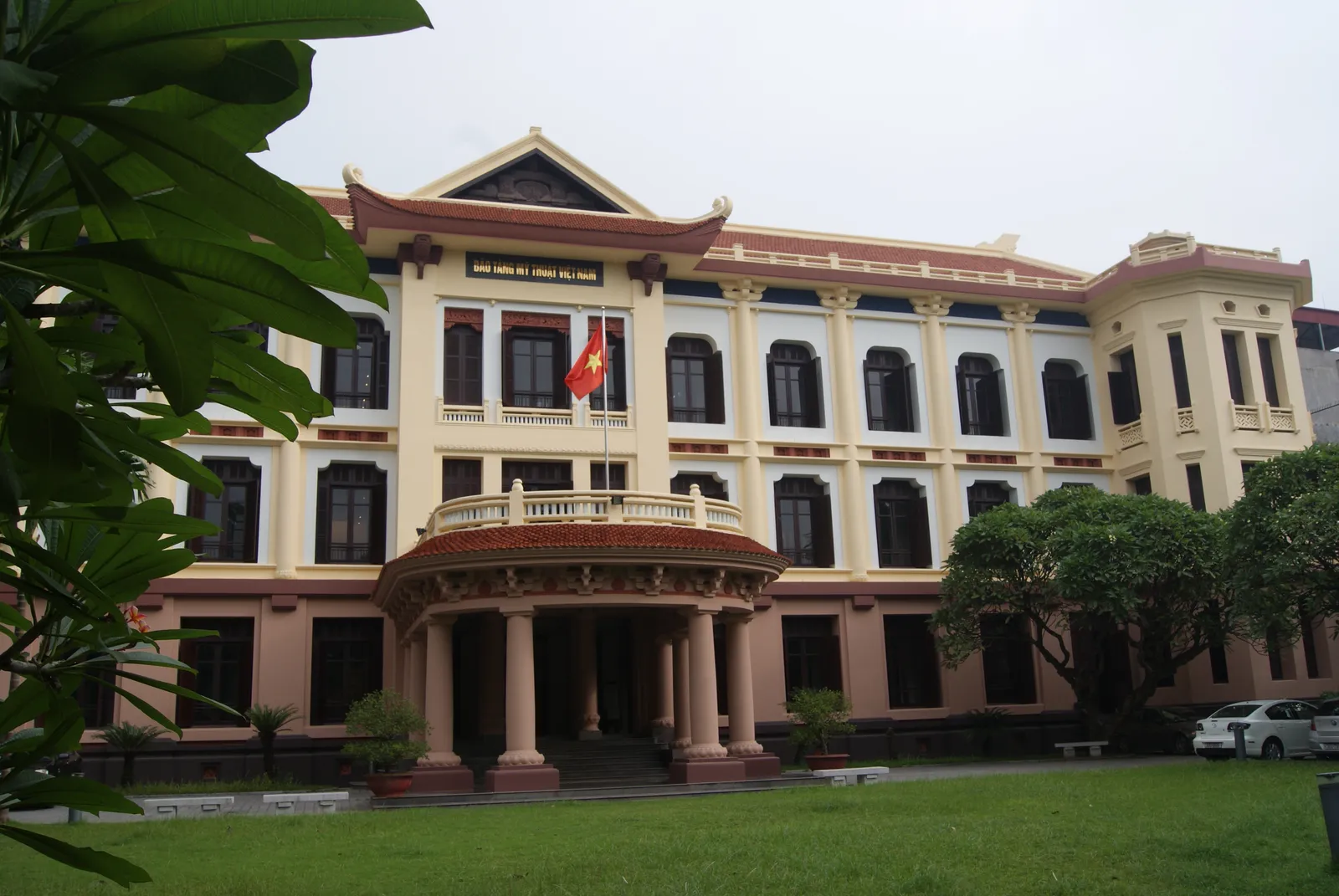 The Vietnam National Museum of Fine Arts in Hanoi, a three-storey cream and pink colonial building with shuttered windows and a tiled Vietnamese roof added on top, seen across its front lawn
