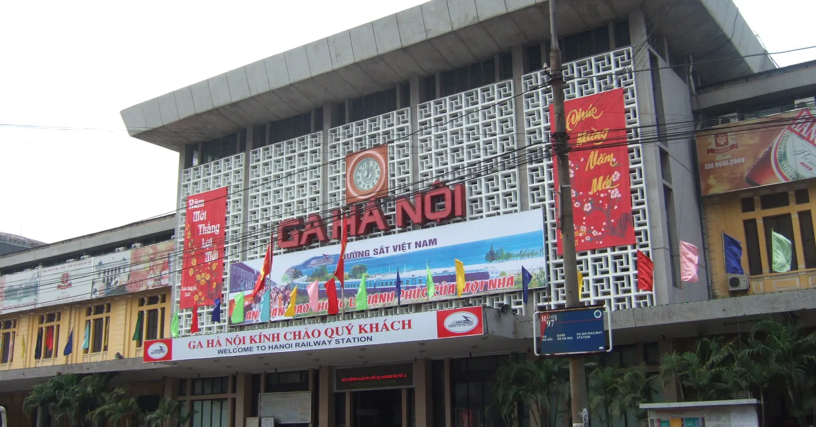 The front of Hanoi Railway Station on Le Duan street, a grey concrete central block with a clock and the words Ga Ha Noi across it, an older yellow colonial wing visible at the left