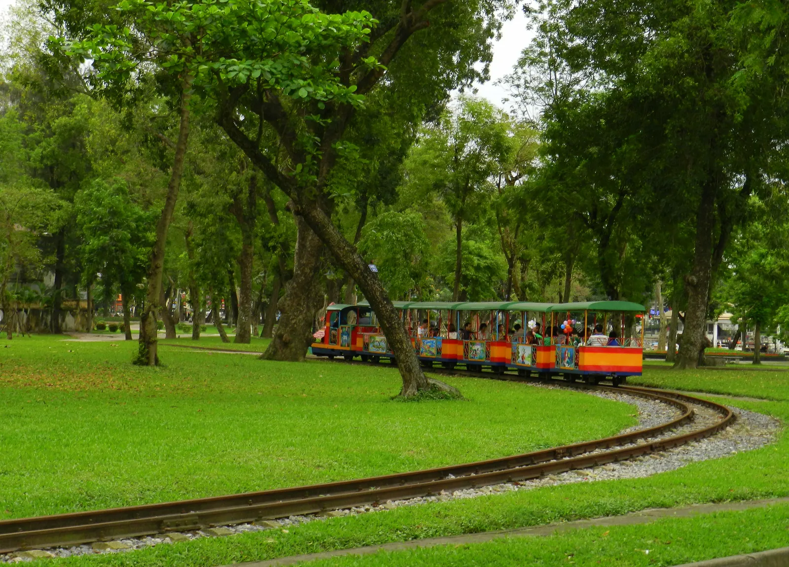 A small open-sided tourist train curving along a narrow track through mown grass and mature trees in Thong Nhat Park in Hanoi