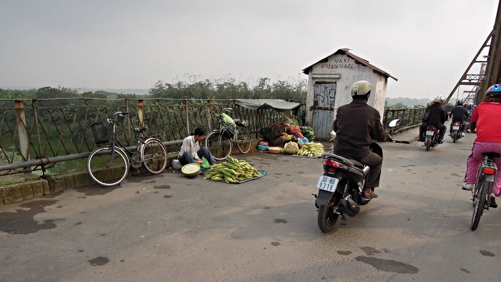 A seller squatting on the walkway of Long Bien Bridge in Hanoi behind bunches of green bananas laid on the ground, with motorbikes and a cyclist passing along the lane beside her
