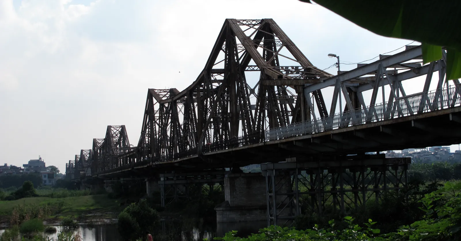 Long Bien Bridge in Hanoi seen from the riverbank, a long line of dark steel trusses on concrete piers with one squarer replacement span among the original diamond-shaped ones