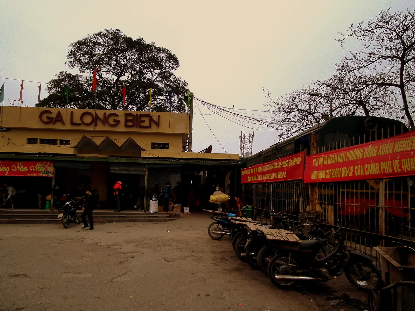 The frontage of Long Bien railway station in Hanoi, a low yellow building with GA LONG BIEN in red lettering on a sign above the entrance, motorbikes parked along the forecourt