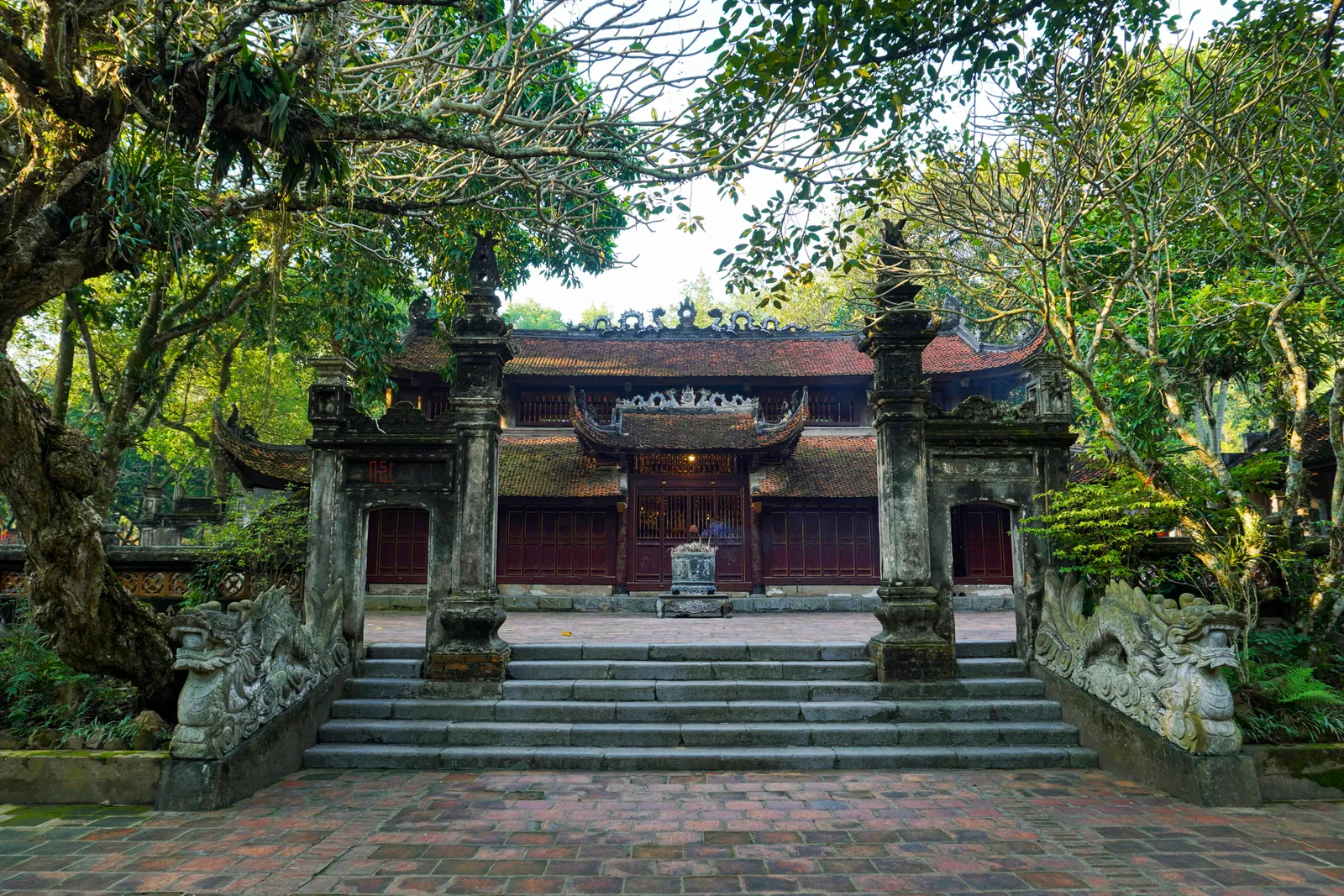 The main hall of Soc Temple in Soc Son, a dark-timbered building with a tiled roof behind two carved stone pillars, reached by a broad flight of steps under old trees