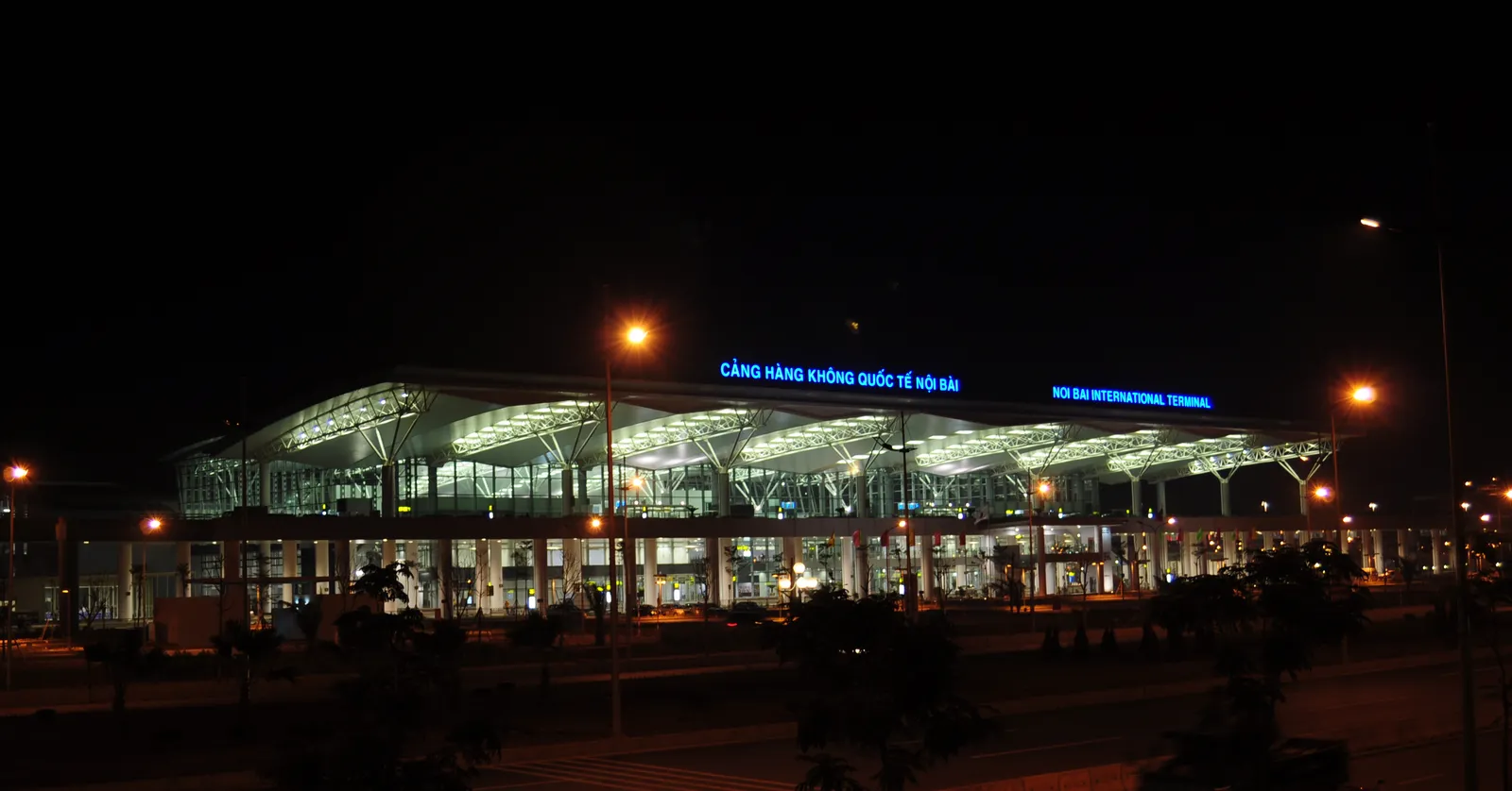 The international terminal at Hanoi's Noi Bai airport lit up after dark, its long glass frontage glowing and the airport's name spelled out in Vietnamese and English across the roofline